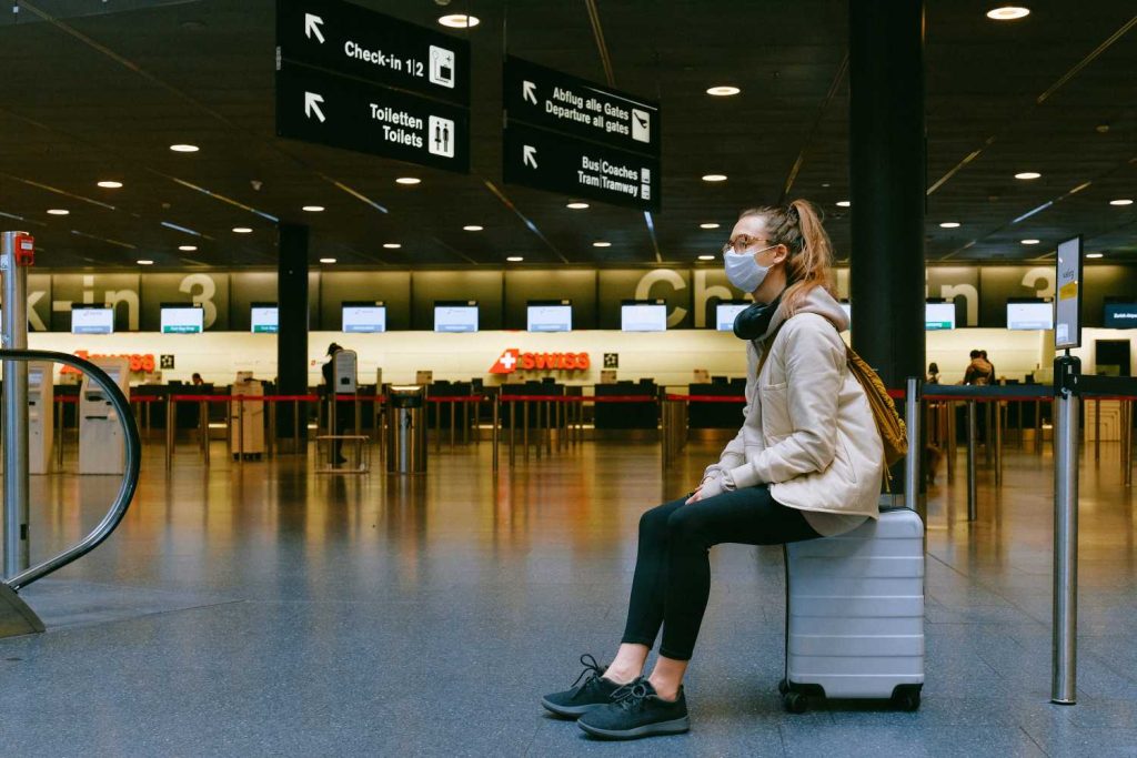 A woman wearing a mask sits on her suitcase in an airport