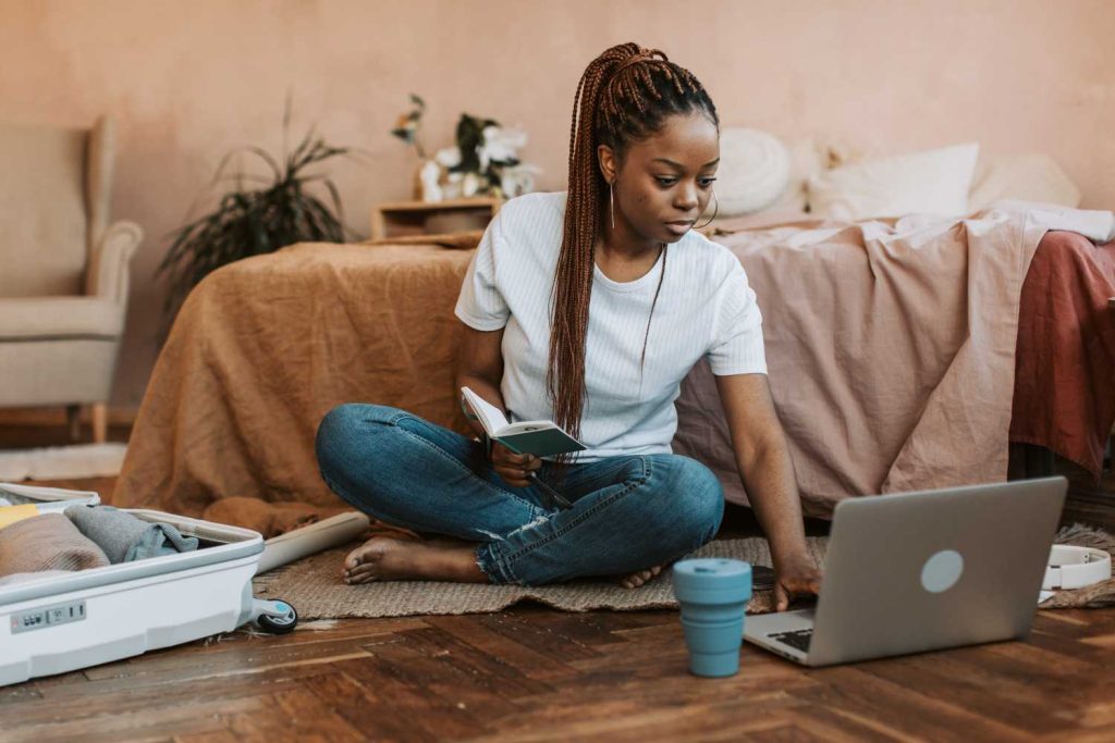 A woman sits on the floor of her bedroom next to a partially-packed suitcase and open laptop