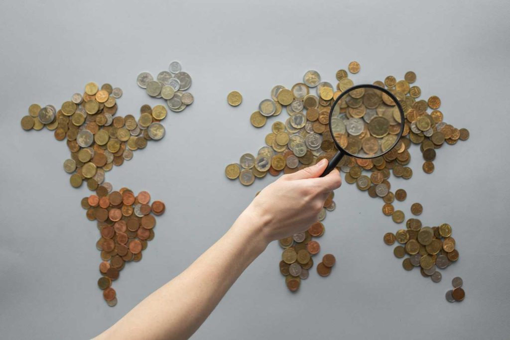 A pile of coins is spread out in the shape of a world map, with a magnifying glass held over it.