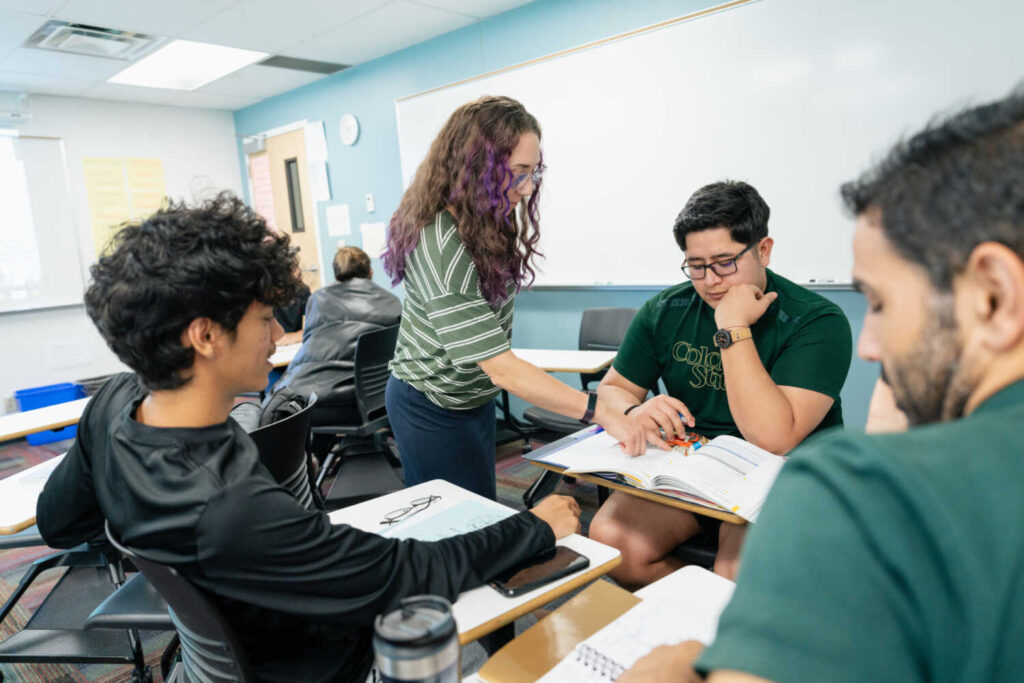 Students sitting in a circle in the classroom with books open. Instructor standing and pointing to a text.