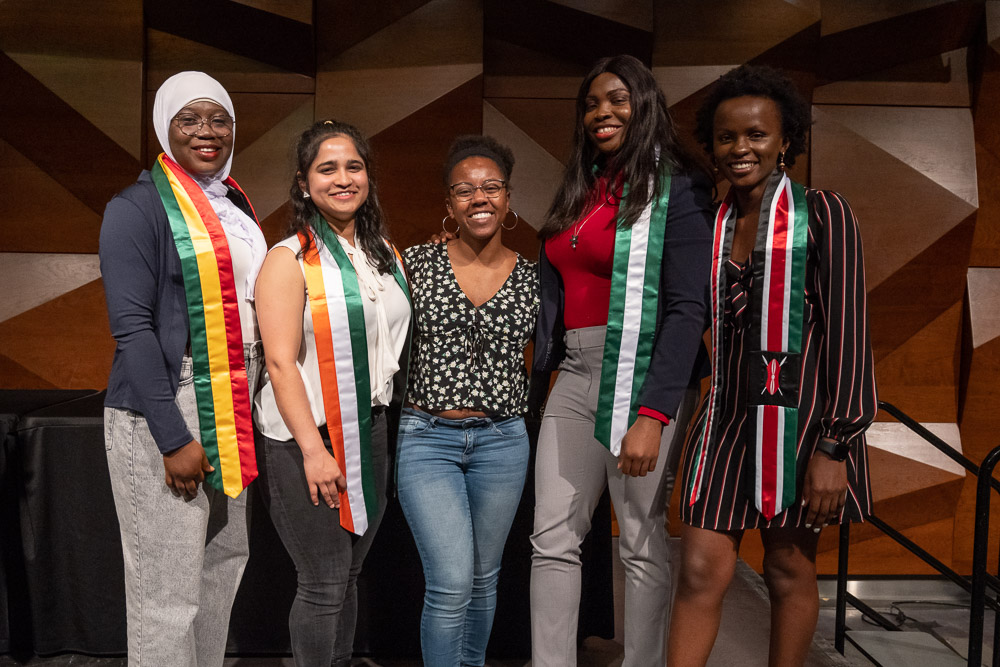 A group of 5 students smile on stage wearing stoles that represent the flags from their home countries.