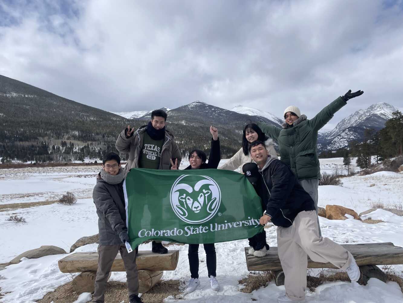 A group of International Students smiling with a Colorado State Flag in front of snowy mountains