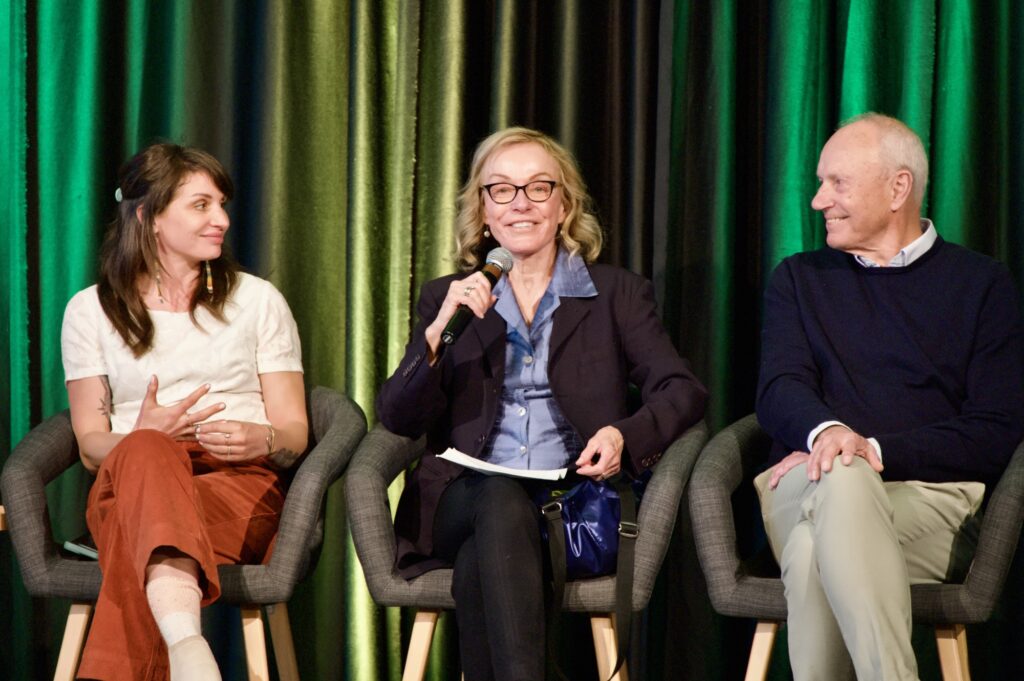 Three panelists sit on a stage, the individual in the middle holds a microphone and is smiling.