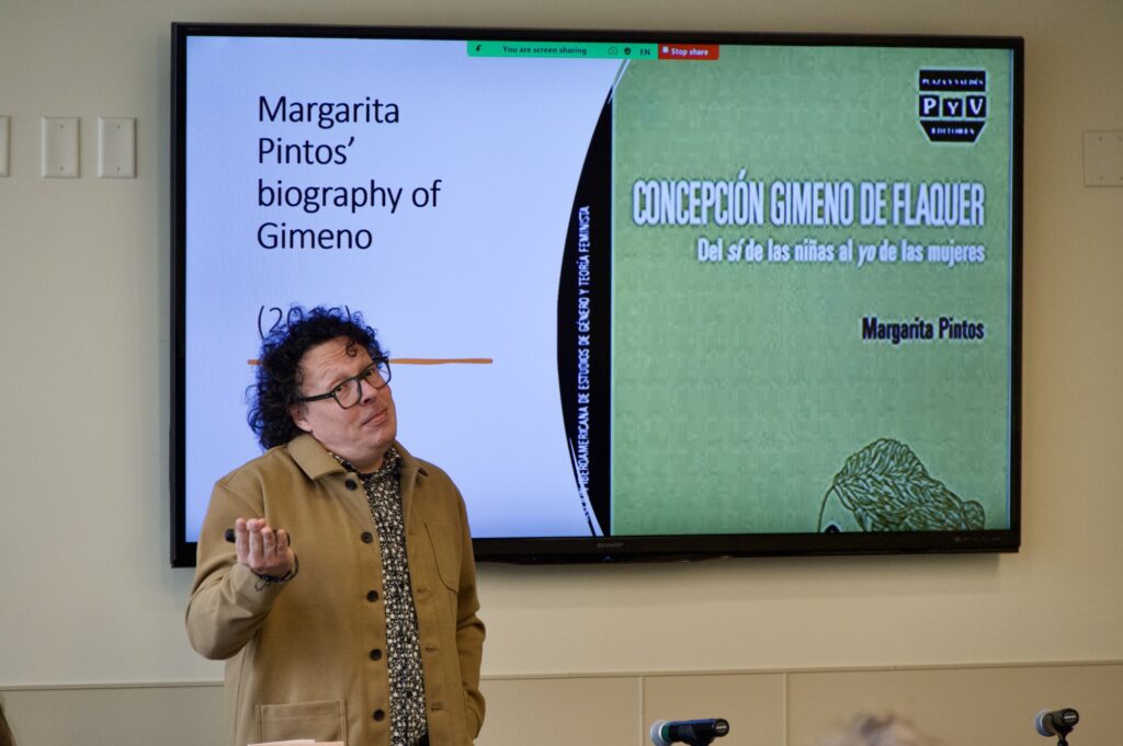 A speaker holds up one arm in a shrug as they stand before their presentation slides at the 2025 International Symposium.