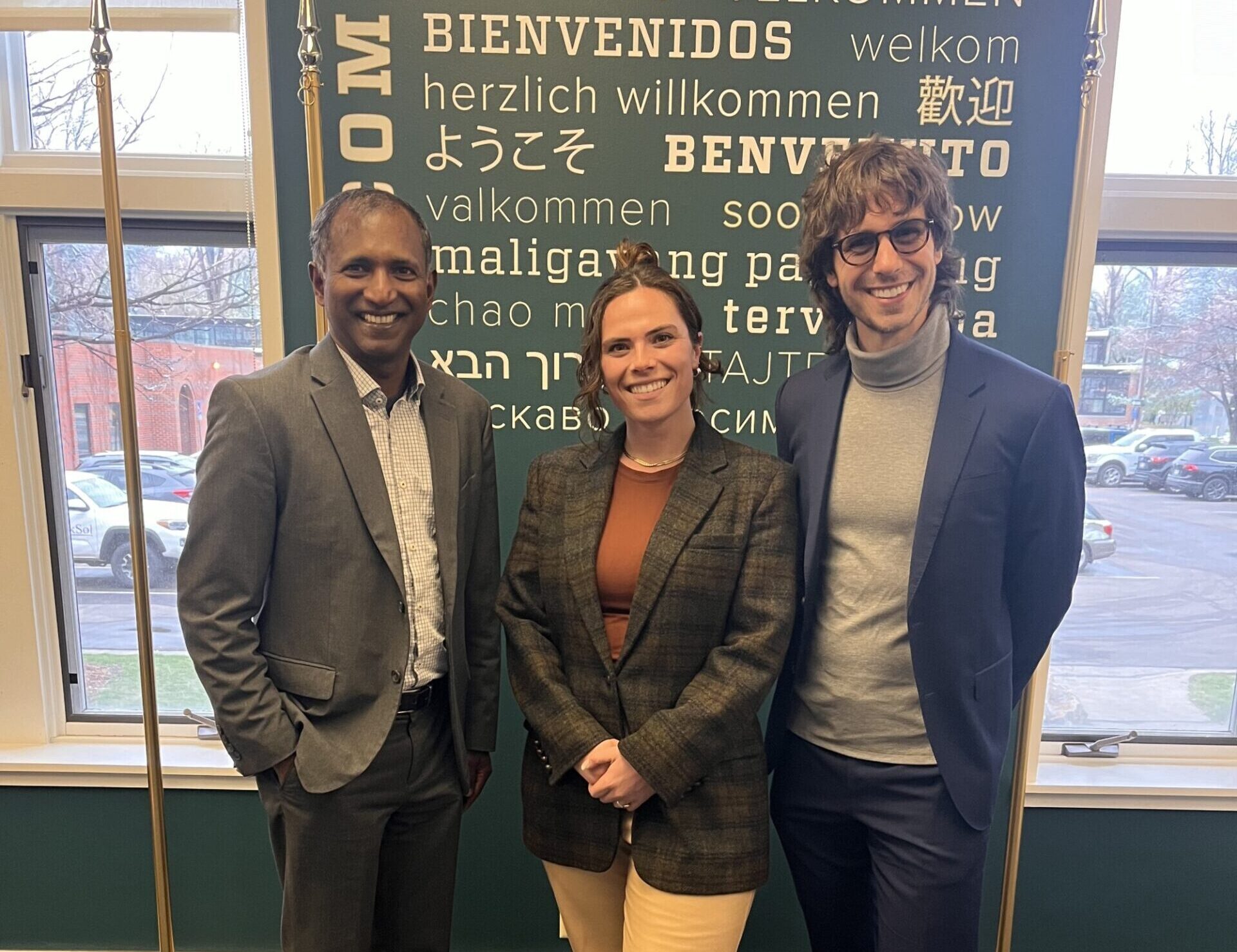 Three individuals smiling in front of a wall that says welcome in several languages