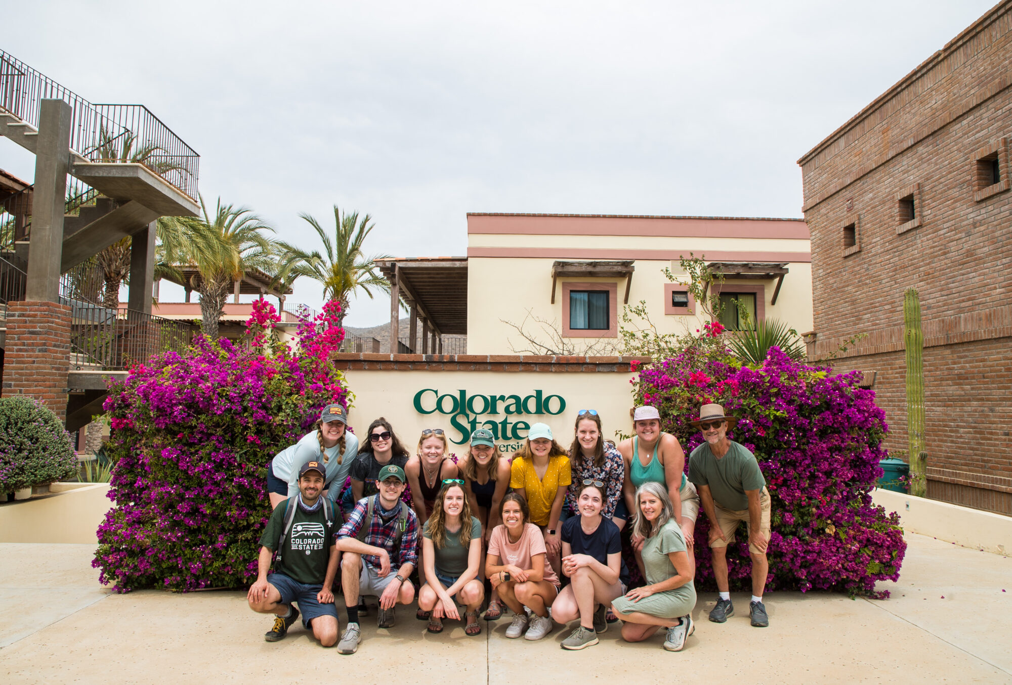 A group of students smiling in front of a Colorado State University sign with palm trees and purple flowers in the background