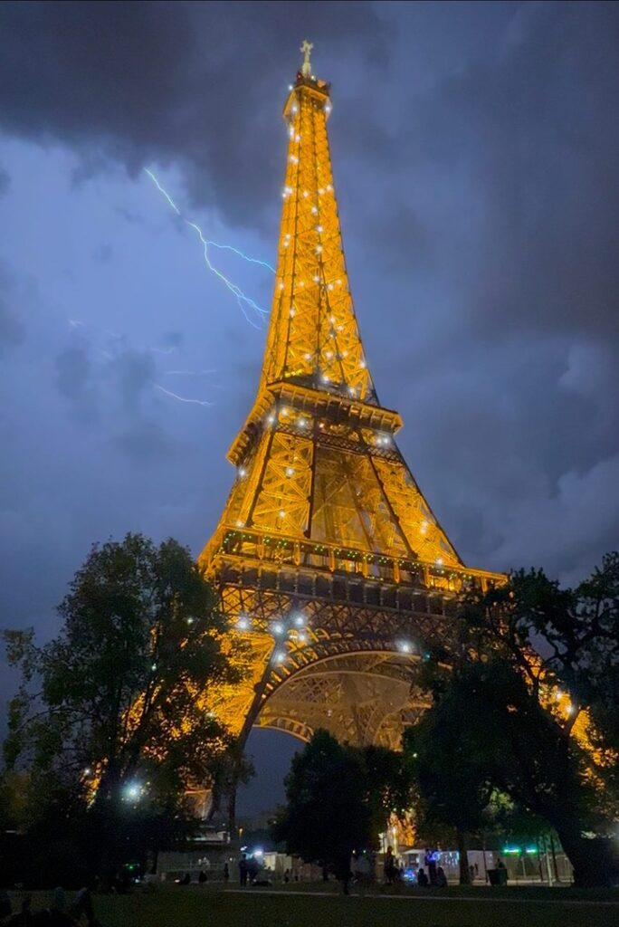 The Eiffel Tower is illuminated on a stormy night as lightning flashes in the background.