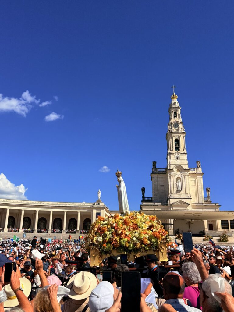 A large densely packed crowd of people stand in the bright sunshine looking up at a statue of the Virgin Mary in front of a large church. At the base of the statue is a small mound of yellow and orange flowers.