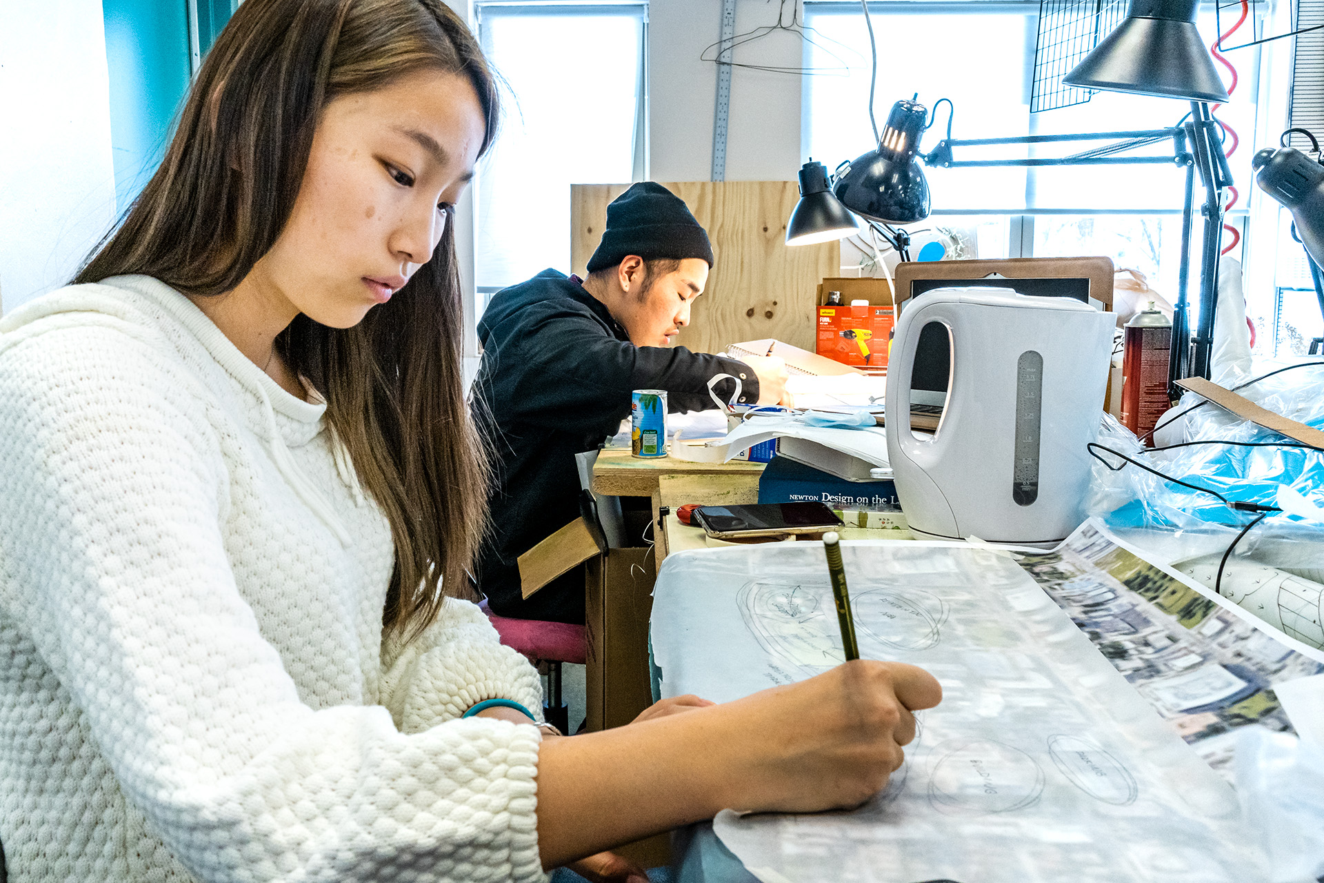 In the foreground, a student writes on a piece of paper while another student works on a project in the background