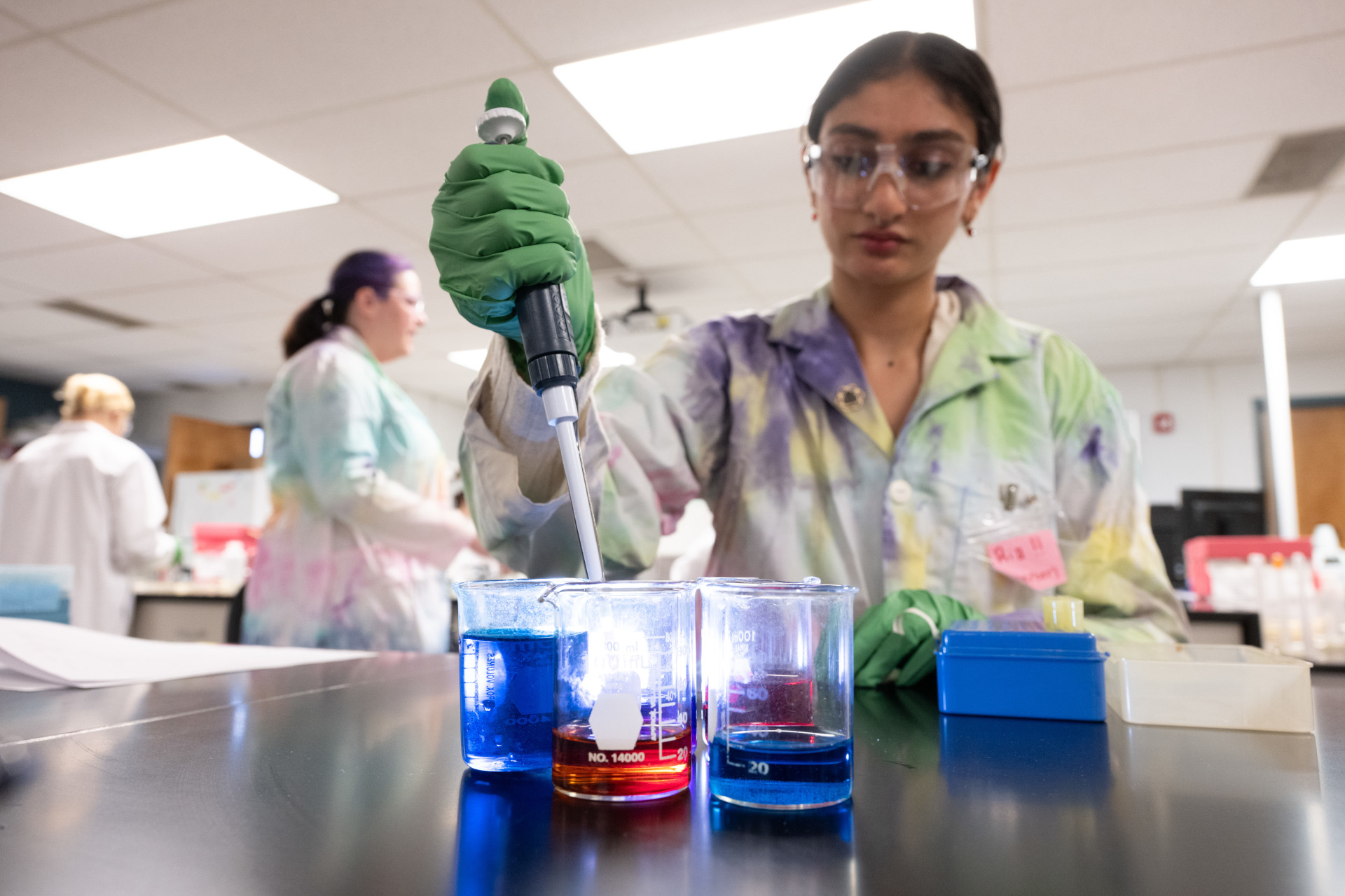 A student wearing a lab coat and goggles performs a science task in a lab