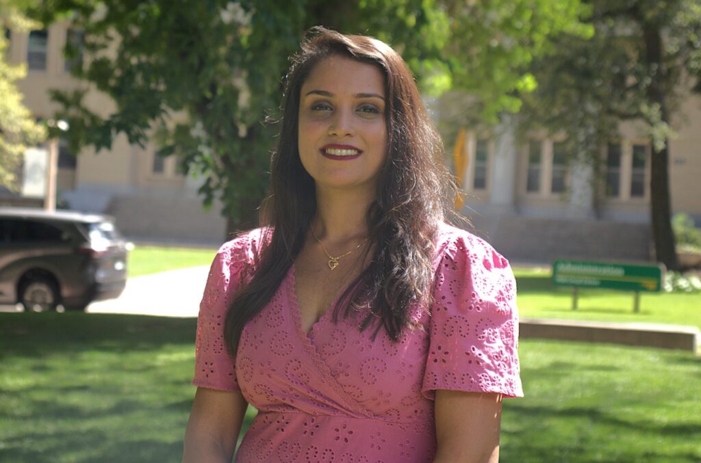 A headshot photo of a woman wearing a pink shirt in front of trees on a sunny day