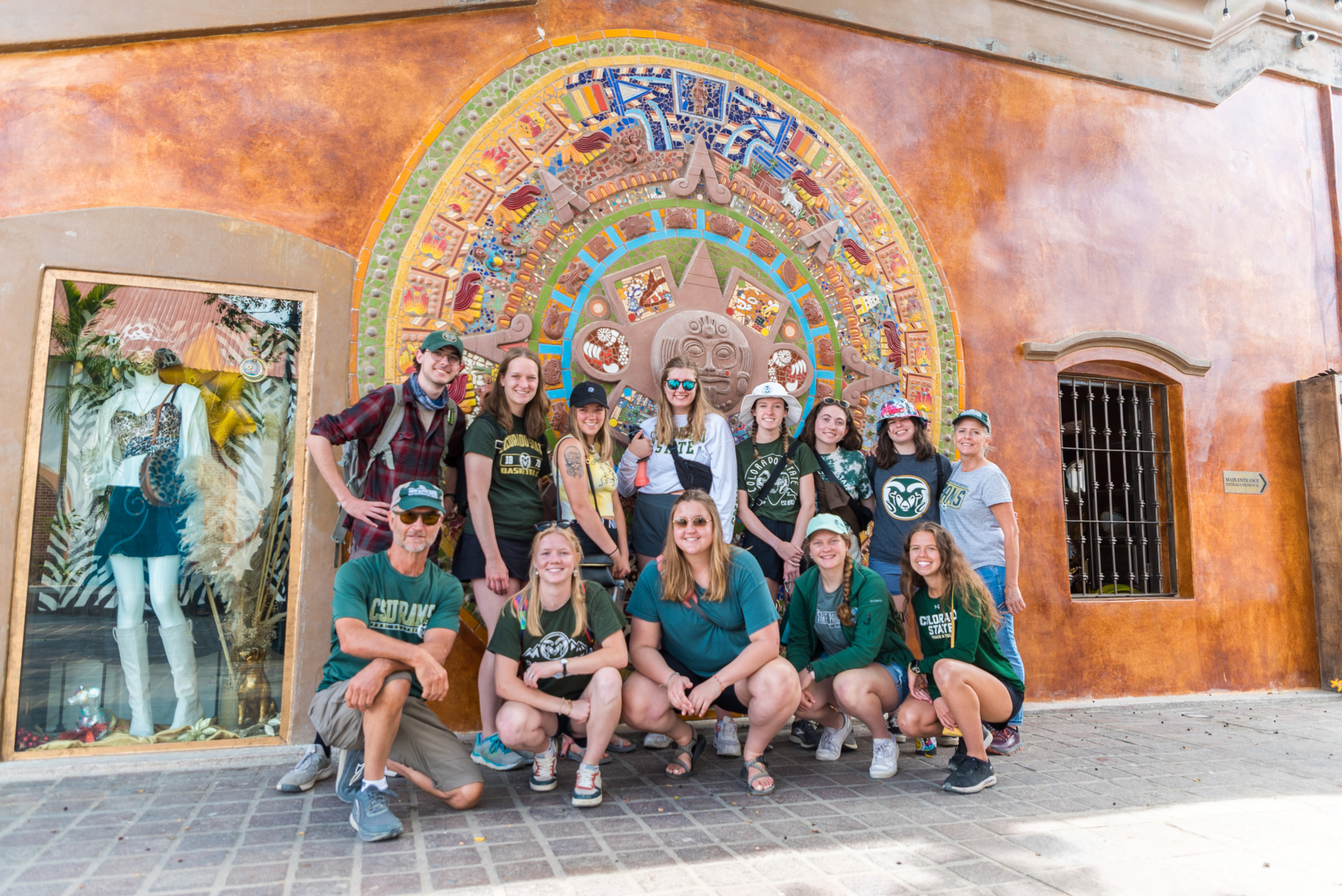 A group of students in CSU gear pose in front of an orange mosaic wall
