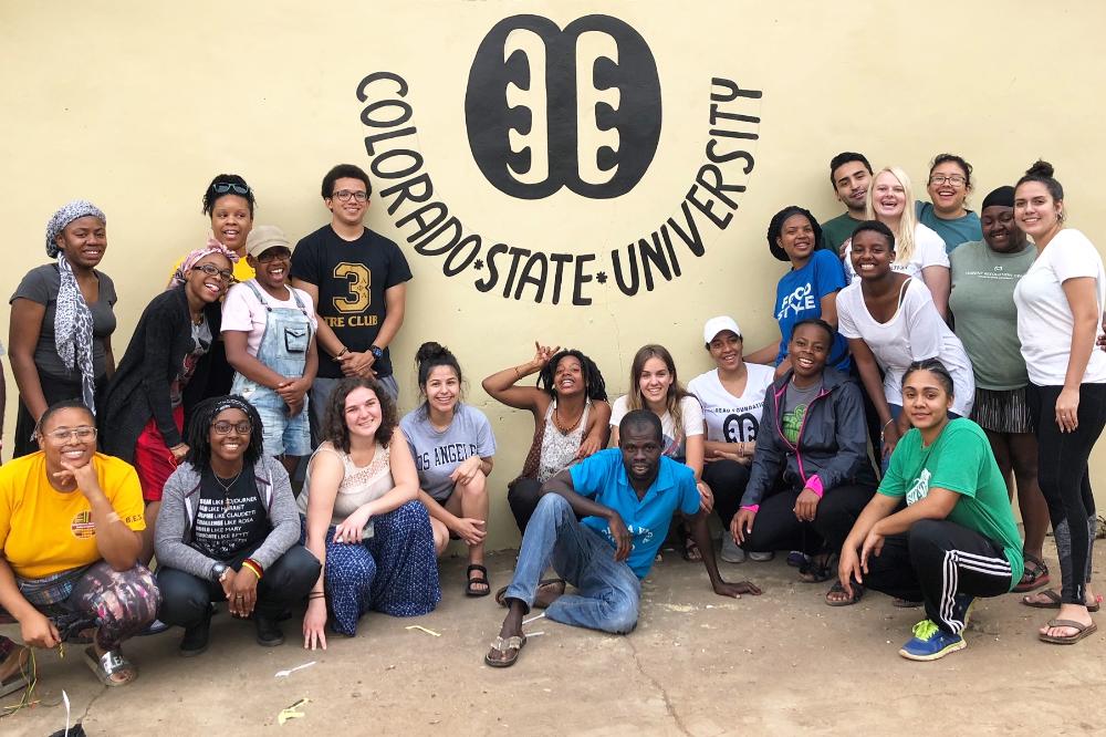 A group smiles in front of a painting on a wall that says Colorado State University