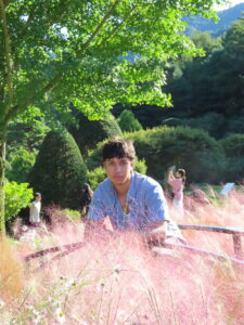 A student sits in tall grass with a manicured garden in the background