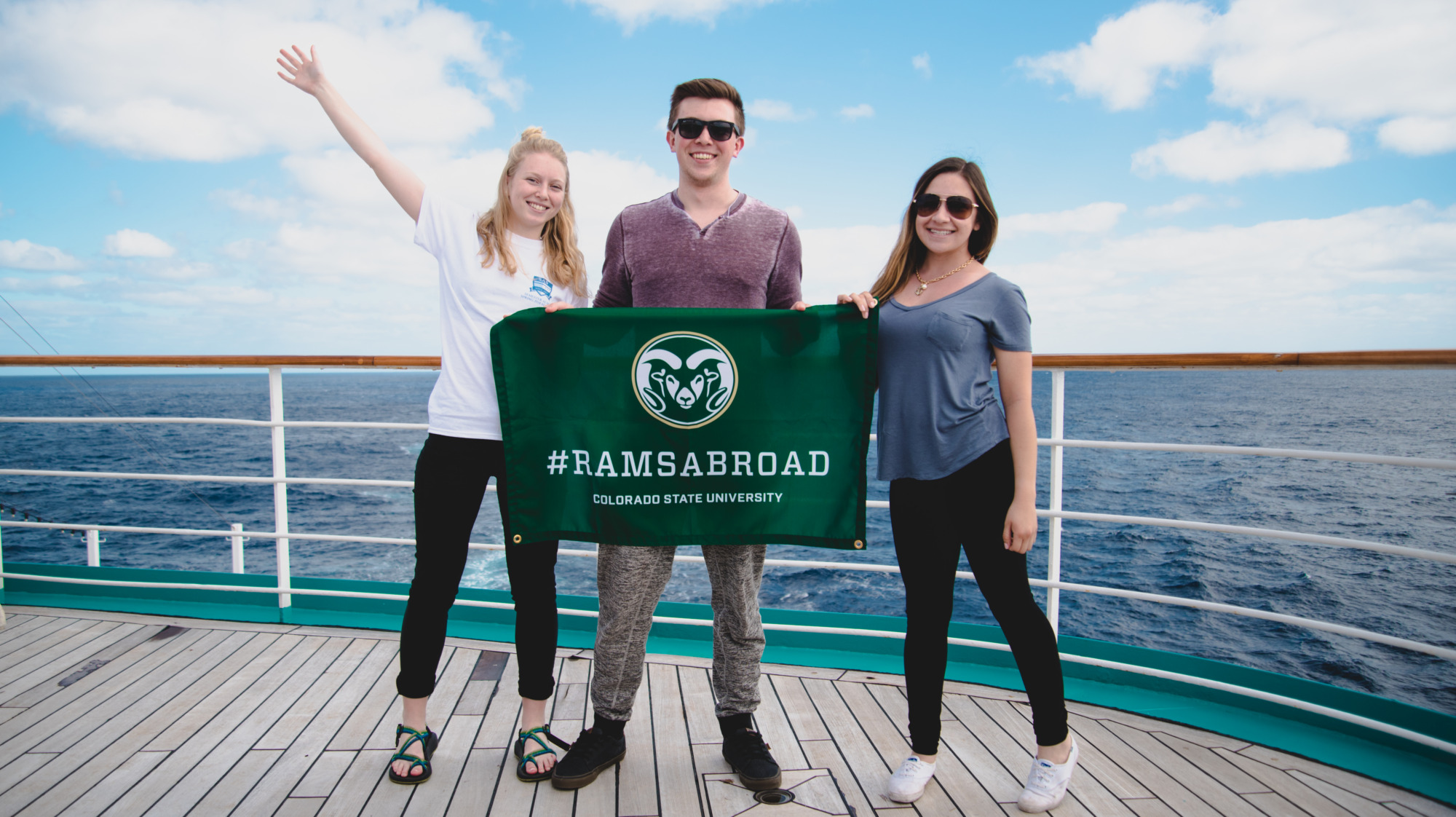 3 students holding a Rams Abroad Flag on the back of a ship