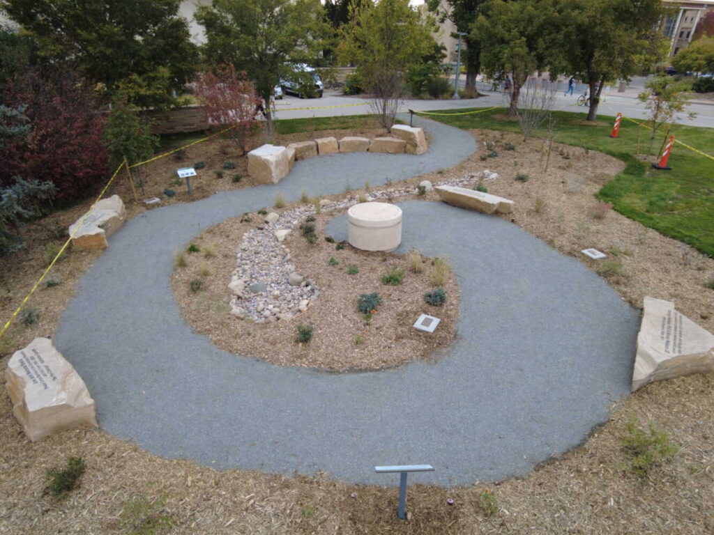 A garden with a spiral path leading to a stone structure in the center. Sitting stones surround the path. 