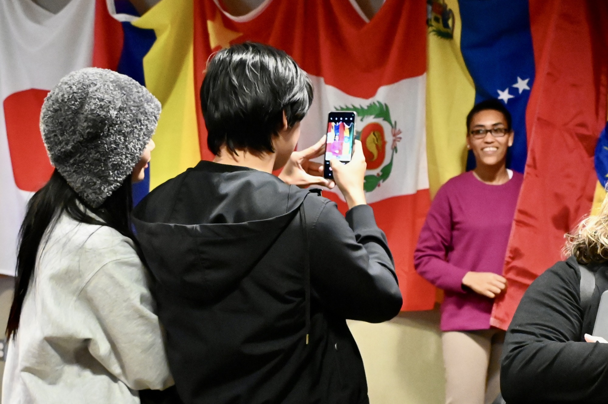 Two students taking a photo of a woman in front of a wall of flags from around the world